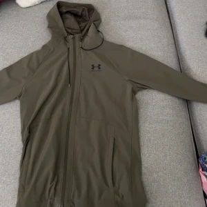 Under Armour vindjacka i beige - Säljer en beige vindjacka från Under Armour med huva och dragkedja fram. Jackan är tillverkad i ett lätt, vattenavvisande material och har en modern, sportig passform. Den har en diskret logga på bröstet och är perfekt för aktiva dagar i blåst och regn. En stilren och funktionell jacka för den aktiva personen.