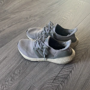 Grå Adidas Ultraboost sneakers - Snygga grå Adidas Ultraboost sneakers med stickad ovandel och vita Boost-sulor. Skorna har snörning och diskreta plastdetaljer på sidorna. Perfekta för dig som gillar sportig stil och vill ha bekväma skor till vardags. Defecter är hälarna i båda skor samt höger snöre där liten plastande saknas som ses på bild 4-5. Skick 7-10 därför annars bra träningssko och väldigt bekväm!