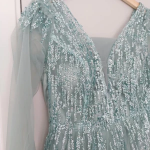 Ljusblå glittrig långklänning - Magisk ljusblå långklänning med transparenta långa ärmar och massor av glittrande paljetter upptill. Klänningen har v-ringning och ett luftigt, voluminöst kjolparti i tyll. Perfekt för bal eller fest där du vill sticka ut.