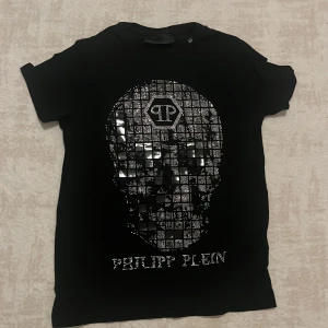 Svart Philipp Plein t-shirt med strass - Svart t-shirt från Philipp Plein med korta ärmar och rund hals. Framsidan har ett stort tryck och logga i glittrande strass. Baksidan har en metallisk PP-logga vid nacken. Snygg och edgy design som sticker ut.
