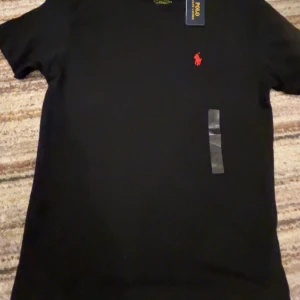 Svart t-shirt Polo Ralph Lauren - Svart t-shirt från Polo Ralph Lauren med röd broderad logga på bröstet. Modellen är custom slim fit och har korta ärmar. Klassisk rund halsringning och mjukt bomullsmaterial. Perfekt för en clean och stilren look.