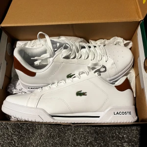 Vita sneakers från Lacoste - Fräscha vita sneakers från Lacoste med bruna detaljer vid hälen och klassisk krokodil-logga på sidan. Skorna har snörning, perforerad tå och är tillverkade i slätt syntetmaterial. Perfekta för en clean och sportig look.