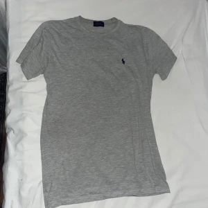 Grå t-shirt från Polo Ralph Lauren - Snygg grå t-shirt från Polo Ralph Lauren med klassisk rund hals och korta ärmar. T-shirten har en liten mörkblå broderad logga på bröstet och är gjord i mjuk bomull. Perfekt basic-plagg med stilren vibe.
