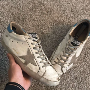 Golden Goose vita sneakers med stjärna - Golden Goose sneakers i vitt skinn med grå mockadetaljer och ikonisk stjärna på sidan. Skorna har grå snörning, blå hälkappa och GGDB-logga på plösen. Snygg vintage-look och platt sula i gummi. Strl 44. Skick: 7,5/10 fräscha, endast lite slitna här och där. Nypris runt 6000kr. Hör av er för mer information osv!