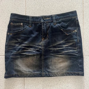 Mörk jeanskjol med kedja och nitar - Cool mörkblå jeanskjol från Sweet med slitningar, silverfärgade nitar och kedjedetalj vid fickan. Fickorna har glittrande stenar och paljetter för extra bling. Klassisk femficksmodell med kontrastsömmar och knappstängning framtill. Strl M men väldigt liten i storleken så den passar som s/xs