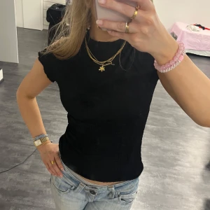 Svart ribbad topp med volangkant - Supersnygg svart topp med ribbad struktur och liten volangkant vid halsen. Modellen är figurnära med korta ärmar och lätt stretchigt material. Perfekt till jeans eller kjol för en clean look.