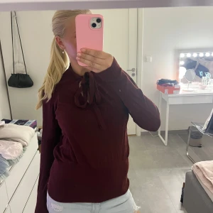 Vinröd stickad tröja med snörning - Mysig vinröd tröja med långa ärmar och snörning i halsen. Tröjan är stickad och har en snygg v-ringning när snörningen är öppen. Perfekt för höst och vinter när du vill vara både bekväm och stilren.