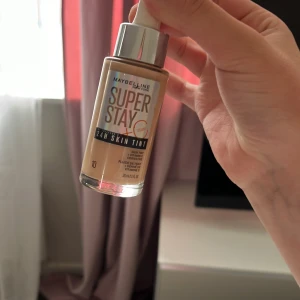 Maybelline Super Stay Skin Tint 10 - Maybelline Super Stay 24H Skin Tint i nyansen 10. Flytande foundation med vitamin C som ger en naturlig finish och håller hela dagen. Kommer i en glasflaska med silvrig pump och lock. Perfekt för dig som vill ha en lätt täckning med glow.