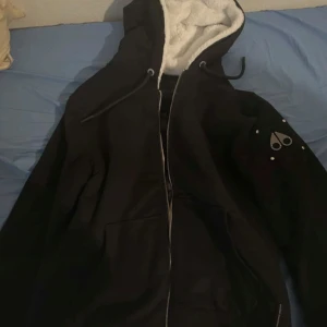 Svart hoodiejacka Moose Knuckles - Svart hoodiejacka från Moose Knuckles med dragkedja, snörning i huvan och mjukt vitt foder. Jackan har deras ikoniska logga i metall på ärmen och stora fickor framtill. Perfekt för dig som gillar streetwear och vill ha något både snyggt och bekvämt.