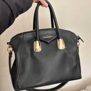 Svart Givenchy handväska i skinn - Säljer en svart handväska från Givenchy i slätt skinn med guldiga metalldetaljer och dragkedja. Väskan har både handtag och axelrem, vilket gör den enkel att bära på olika sätt. Klassisk och stilren design med Givenchy-logga framtill.