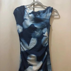 Blommig blå långklänning - Säljer en långklänning med blommigt mönster i olika blå och vita nyanser. Klänningen har en figurnära passform och är ärmlös med rundad hals. Materialet känns mjukt och följsamt, perfekt för dig som vill sticka ut med en cool och unik look.