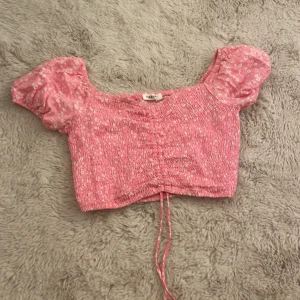 Rosa blommig croppad topp Terranova - Supersöt rosa croppad topp från Terranova med små vita blommor, puffiga korta ärmar och smockad kropp. Toppen har snörning framtill för extra detalj och en feminin v-ringning. Perfekt för dig som gillar romantisk och trendig stil.