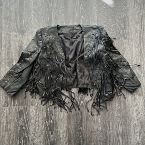 Svart skinnjacka med fransar H&M - Cool svart skinnjacka från H&M med långa fransar framtill och på ärmarna. Jackan har snygga snörningsdetaljer och metallöljetter längs axlar och ärmar. Skadad på axeln därav priset 