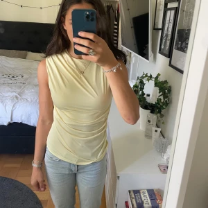 Ljusgul ärmlös topp från H&M - Säljer en ljusgul ärmlös topp från H&M med draperad effekt över axel och sida. Aldrig använd och prislappen är kvar.