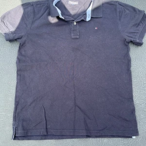 Mörkblå pikétröja Tommy Hilfiger - Snygg mörkblå pikétröja från Tommy Hilfiger med klassisk krage och två knappar framtill. Tröjan har en diskret logga broderad på bröstet och ljusblå detaljer på insidan av kragen. Perfekt för dig som gillar stilrena och tidlösa plagg.