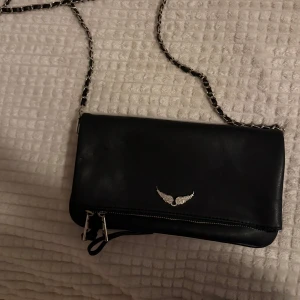 Svart zadig &voltaier axelväska passar bra till skolan  - Snygg svart axelväska i skinn med silverfärgad kedja som axelrem och ett glittrigt vingemblem på framsidan. Väskan har dragkedja och får plats med mycket saker. Passar perfekt till skolan eller när man ska ut