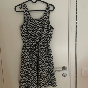 Svartvit mönstrad klänning Divided - Säljer en svartvit kortklänning från Divided med grafiskt mönster. Klänningen är ärmlös och har en rundad halsringning samt en snygg öppen rygg med knyt i nacken. Midjan är lätt rynkad för en skön passform. Perfekt för dig som gillar statement-plagg!