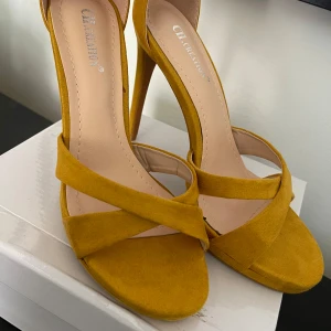 Gula höga klackar från CH Creation - Snygga gula sandaler med hög klack från CH Creation. Skorna har en öppen tå, breda korslagda remmar och en smal ankelrem med spänne. Tillverkade i mjuk mocka som ger en lyxig känsla och passar perfekt till att lyfta din outfit.