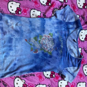 Blå Ed Hardy t-shirt med blommor - Säljer en blå tie-dye t-shirt från Ed Hardy med kort ärm. Framsidan har ett stort blommotiv med strassdetaljer och texten ed Hardy. Tryckt Ed Hardy-logga i gult både fram och bak. Perfekt för dig som gillar färg och statement-plagg. Storleken är egentligen xxl men är snarare mer i storlek S