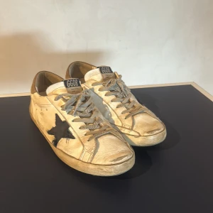 Golden Goose vita sneakers med svart stjärna - Golden Goose sneakers i vitt skinn med ikonisk svart stjärna på sidan och beige detaljer vid hälen. Skorna har snörning med GGDB-tryck och en klassisk rund tå. Perfekta för dig som gillar en avslappnad och trendig streetstyle.
