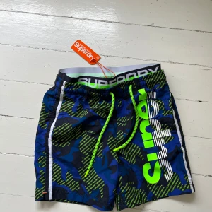 Superdry badshorts helt nya - Snygga badshorts från Superdry i blått och mörkblått camouflagemönster med neongröna detaljer och stor logga på sidan. Shortsen har snörning i midjan, meshfoder och dragkedja på bakfickan. Perfekta för stranden eller poolen.
