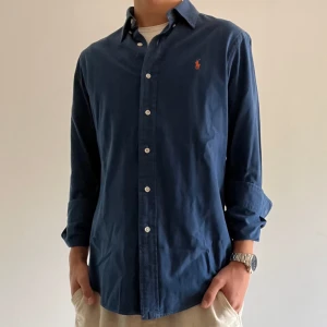 Mörkblå skjorta från Ralph Lauren S - Klassisk mörkblå skjorta från Ralph Lauren i bomull med custom fit. Skjortan har långa ärmar, knappar framtill och den ikoniska broderade loggan i orange på bröstet. Perfekt för en clean och stilren look.