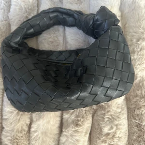 Svart axelväska bottega veneta look alike  - Säljer en svart handväska. Väskan har ett flätat mönster och guldig dragkedja med märkets logga. Den är mjuk och har en cool, modern look som sticker ut. Perfekt för dig som gillar lyxiga detaljer och stilren design. Köptes för 2500kr💞❣️💕