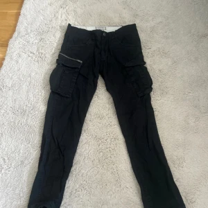 Svarta cargopants från Jack & Jones - Svarta cargopants från Jack & Jones med flera stora fickor på sidorna, dragkedjedetalj och snörning vid benslut. Byxorna har en loose anti fit-passform och är tillverkade i bomull. Perfekta för en avslappnad streetstil.