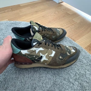 Valentino Rockrunner camo sneakers - Säljer ett par Valentino Rockrunner sneakers med camouflagemönster i grönt, brunt, beige och vitt. Skorna har detaljer i mocka och skinn, svarta snören och en platt sula. Baksidan har en cool mintgrön och vinröd touch med nitar. Perfekt för dig som gillar statement-sneakers. Hör av dig vid frågor eller funderingar! 😊