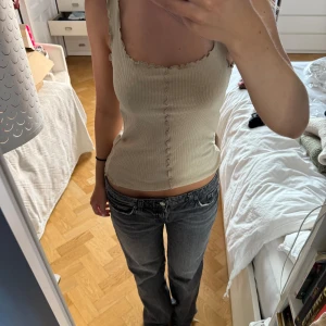 Beige topp - Supersöt beige ribbad linnetopp med vågiga kanter längs axelband, halsringning och nedre kant. Toppen har en figurnära passform och dekorativ söm framtill. Osäker på märke, köpt i Spanien.