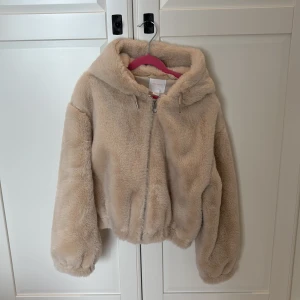 Beige teddyjacka från Gina Tricot - Supermjuk och fluffig beige teddyjacka från Gina Tricot med huva och dragkedja framtill. Jackan har en avslappnad passform och är perfekt för kyliga dagar. Den är helfärgad och har långa ärmar samt elastisk nederkant för extra komfort.