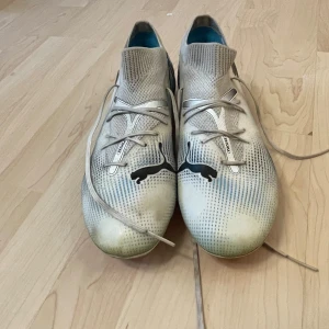 Puma vita fotbollsskor med rosa dobbar utan skolådan  - Säljer ett par vita Puma fotbollsskor med svart logga på sidan och ovansidan. Skorna har snörning, stickad strumpliknande krage och rosa dobbar undertill. Lätta och smidiga för snabba rörelser på planen. Perfekta för dig som vill sticka ut med stil på matchen.