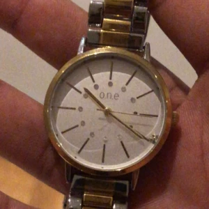 Guld och silver klocka från o.n.e - Snygg klocka från o.n.e med rund urtavla i vitt och detaljer i guld. Armbandet är i metall med både guld- och silverfärgade länkar. Urtavlan har minimalistiska markeringar och små stenar som extra detalj. Perfekt för dig som gillar stilrena accessoarer.