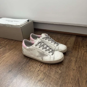 Golden Goose Superstar sneakers vit/rosa - Golden Goose Superstar sneakers i vitt skinn med grå stjärndetalj på sidan och ljusrosa häl. Grå snörning och platt sula. Ikonisk design med coola vintage-vibbar och kontrastfärgade detaljer. Skorna är använda fåtal gånger och har inga deffekter förutom ett litet slitage på hälen på vänster sko, men inget som märks av. Skolåda och dustbag tillkommer.
