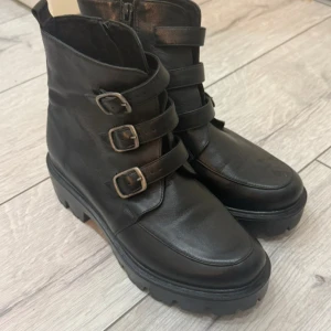 Svarta boots med spännen och dragkedja 100% läder  - Chunky svarta boots i skinn med tre coola spännen framtill och grov sula. Dragkedja på sidan för enkel påtagning. Perfekta för dig som gillar en edgy stil och vill ha skor som sticker ut. Använd 1 gång