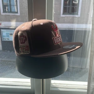 Milwaukee Bucks keps Mitchell & Ness - Snygg brun Milwaukee Bucks keps från Mitchell & Ness med broderade NBA-loggor och detaljer. Fitted modell med platt skärm och tydliga broderier i rött, vitt och beige. Perfekt för dig som gillar basket och streetstyle.