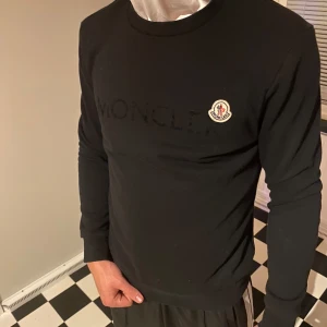 Svart sweatshirt från Moncler - Svart sweatshirt från Moncler med broderad logotyp på bröstet och stort Moncler-tryck framtill. Tröjan har rund hals, långa ärmar och är gjord i mjuk bomull. Perfekt för dig som gillar stilrena och exklusiva märken.