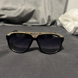 Svarta Burberry solglasögon med gulddetaljer - Snygga svarta solglasögon från Burberry med guldiga detaljer och klassiskt mönster på ovansidan av bågen. Modellen har en cool pilotform och mörka glas. Perfekt accessoar för att lyfta din stil och sticka ut i mängden.