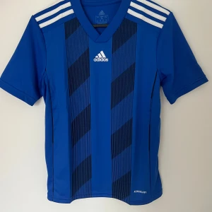 Blå randig Adidas fotbollströja - Snygg blå fotbollströja från Adidas med svarta diagonala ränder och vita detaljer på axlarna. Tröjan har V-ringning, korta ärmar och är gjord i lätt, ventilerande polyester med Aeroready-teknologi. Perfekt för träning eller match!
