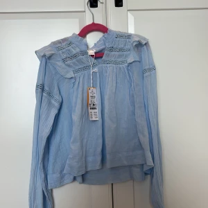 Ljusblå boho blus med spetsdetaljer - Supersöt ljusblå boho blus med volang vid axlarna och spetsdetaljer på bröstet och ärmarna. Blusen har lång ärm och en lätt, luftig känsla som gör den perfekt för vår och sommar. Passar dig som gillar romantisk och chill stil.