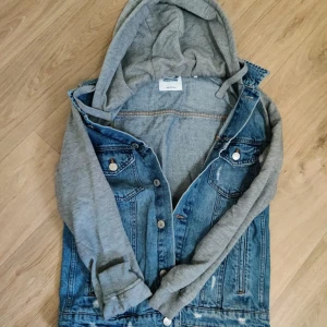 Jeansjacka med grå hoodieärmar - Snygg jeansjacka med grå sweatshirt-ärmar och huva. Klassisk blå denim med knappar framtill och två bröstfickor. Perfekt för en avslappnad streetstyle-look. Jackan har en cool mix av material och passar till det mesta.