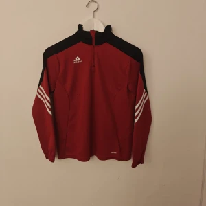Adidas tröja - Snygg röd Adidas tröja med svarta detaljer och vita ränder på ärmarna. Half zip framtill och hög krage ger en sportig vibe. Tillverkad i polyester med Climacool-teknologi som håller dig fräsch. Perfekt för träning eller chill. Långärmad och normal passform.
