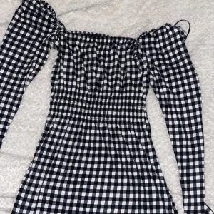 Svartvit rutig klänning H&M Divided - Nyköpt oanvänd!! Supersöt svartvit rutig klänning från H&M Divided i storlek XS. Klänningen har långärmade puffärmar, smockad midja och offshoulder-design. Materialet är mjukt och känns som bomull, perfekt för dig som gillar trendiga och bekväma outfits.