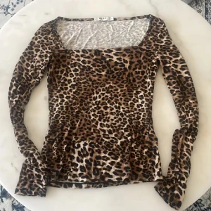 Leopardmönstrad topp från NA-KD - Säljer en snygg långärmad topp från NA-KD i leopardmönster. Modellen har fyrkantig urringning och sitter tight mot kroppen. Färgerna går i brun, beige och svart. Perfekt att styla med jeans eller kjol för en cool look.