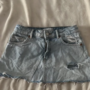 Ljusblå jeanskjol från Zara - Säljer en ljusblå jeanskjol från Zara med slitna detaljer och rå kant längst ner. Kjolen har klassiska fickor fram och bak, bälteshällor och dragkedja med knapp. Perfekt för dig som gillar en chill och trendig streetstil.