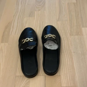 Svarta loafers från Novita med guldkedja - Stilrena svarta loafers från Novita i skinn med chunky guldfärgad kedjedetalj framtill. Platt sula och öppen häl ger en modern look, perfekt till jeans eller kostymbyxor. Klassisk rund tå och enkel att bara slinka i. Riktigt skinn.