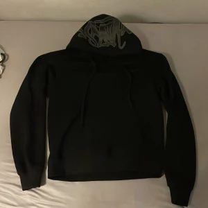Svart hoodie från synaworld - Svart hoodie från synaworld i storlek M. Tröjan har en stor huva med coolt grått tryck och snörning framtill. Materialet är mjukt och känns som bomull, perfekt för chill dagar. Långärmad och med klassisk loose passform.