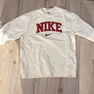 Vit Nike sweatshirt med röd logga - Snygg vit sweatshirt från Nike med stor röd broderad logga och svart swoosh på bröstet. Tröjan har långa ärmar med ribbade muddar och coola röd-svarta detaljer vid ärmsluten. Perfekt för snygg stil.