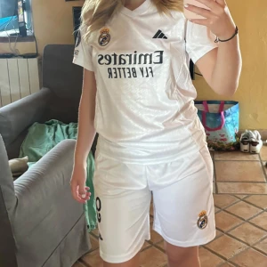 Vit Real Madrid set  - Säljer en vit Real Madrid fotbollströja med klubbmärke och sponsortryck på bröstet. Tröjan är kortärmad och har en klassisk sportig passform, perfekt för dig som älskar fotboll och vill representera ditt favoritlag. Shortsen medföljer! De är i barnstorlek men skulle säga att de passar XS. Nmr 9 mbappe på ryggen