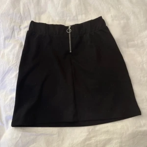 Svart kortkjol med dragkedja - Snygg svart kortkjol med rak passform och en cool silverfärgad dragkedja framtill med ringdetalj. Kjolen har resår i midjan för extra komfort och är gjord i ett mjukt bomullsmaterial. Perfekt att matcha med en oversized hoodie eller en croppad topp.